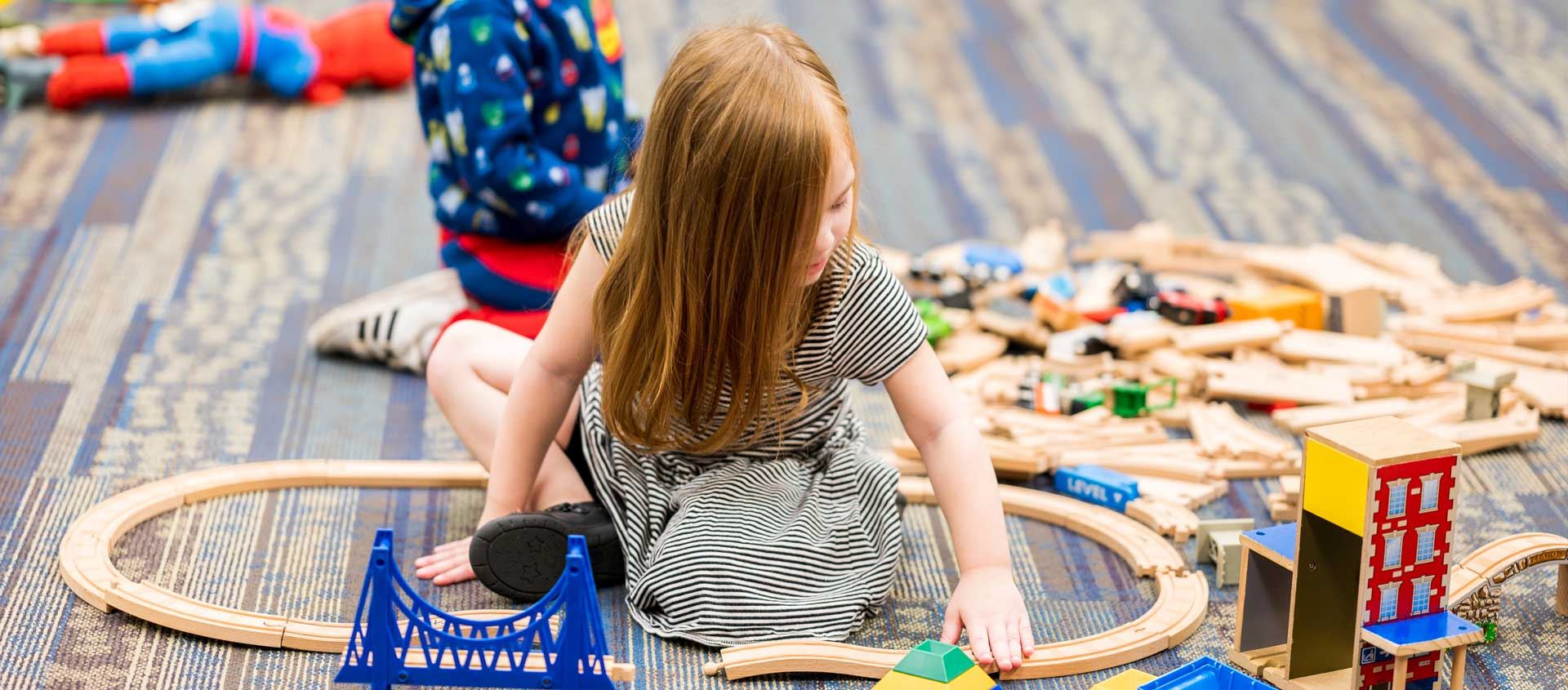 A preschool-aged girl plays with trains while another child sits near her