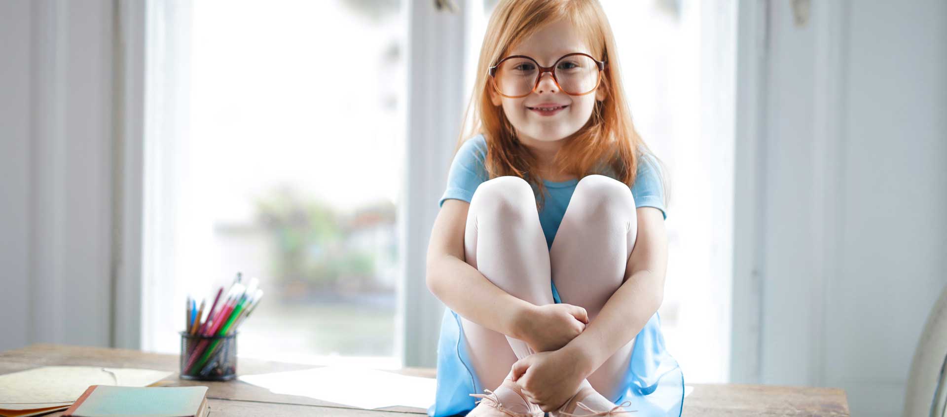 A smiling girl with red hair and glasses sits atop a desk.
