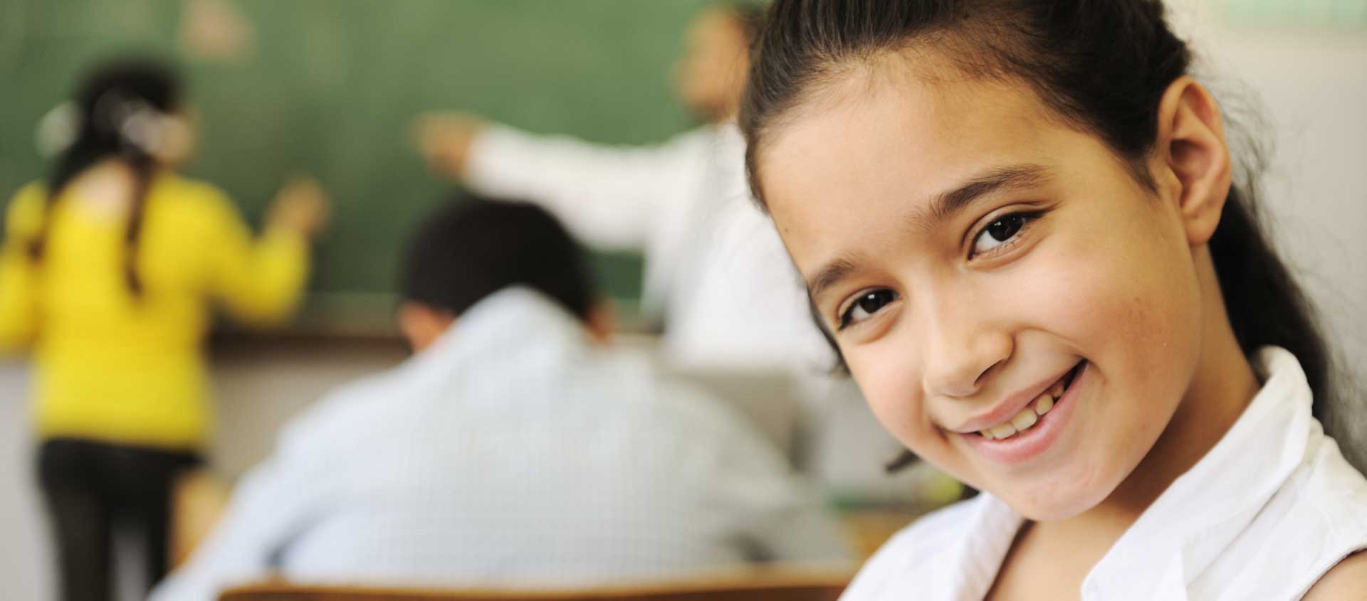A school-aged girl with dark hair smiles ina classroom setting.