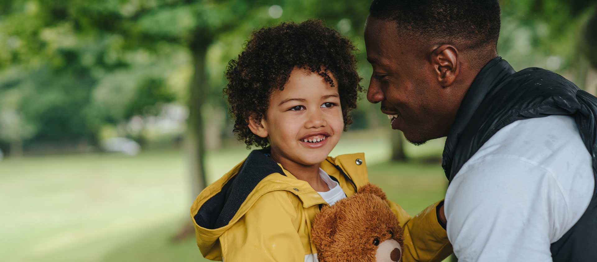 A Black American father and his preschool-aged child embrace in an outdoor setting..