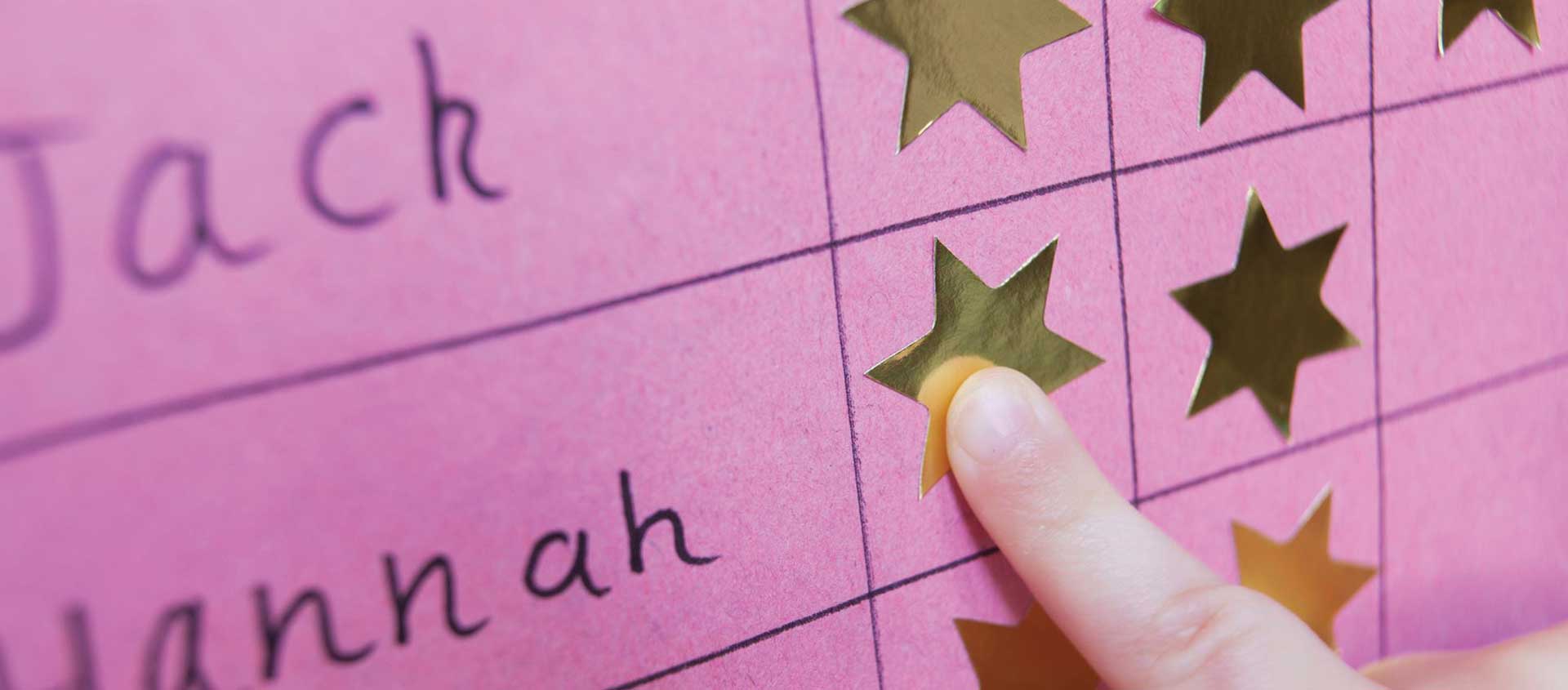 A child's hand, placing a gold star sticker on a pink sticker chart next to the name, "Hannah."