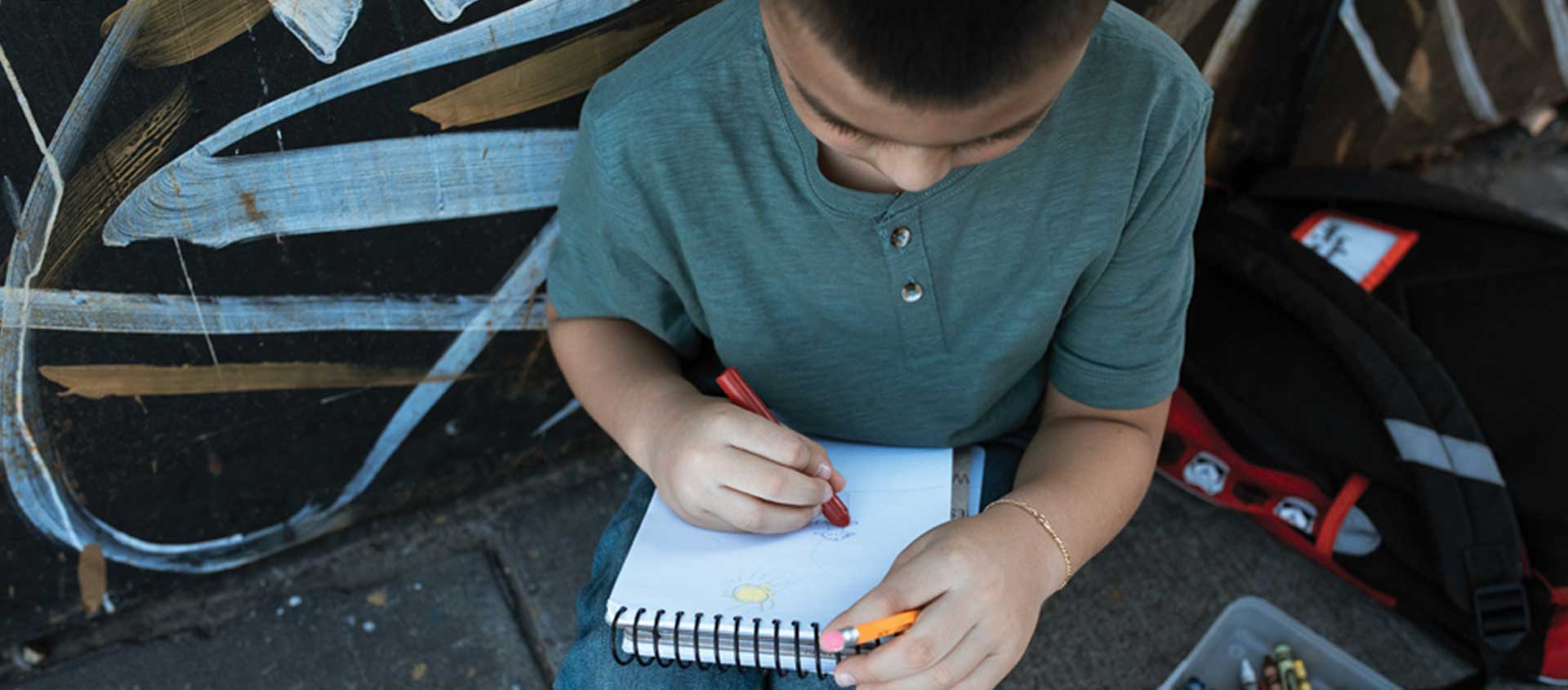 A young boy sits on a sidewalk with a sketch book open on his lap. 