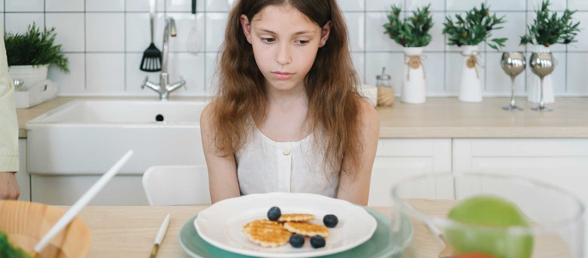 A school-aged girl sits in front of a plate of waffles with a sad look
