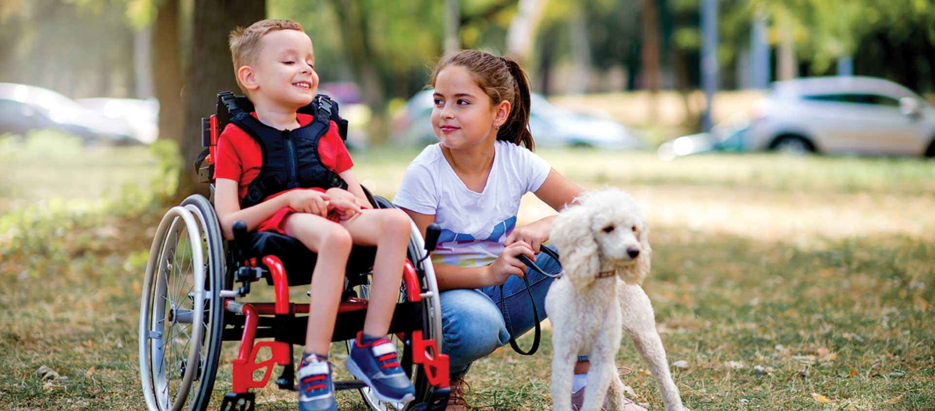 A school-aged Caucasian girl crouches next to a younger Caucasian boy who uses a red wheelchair.