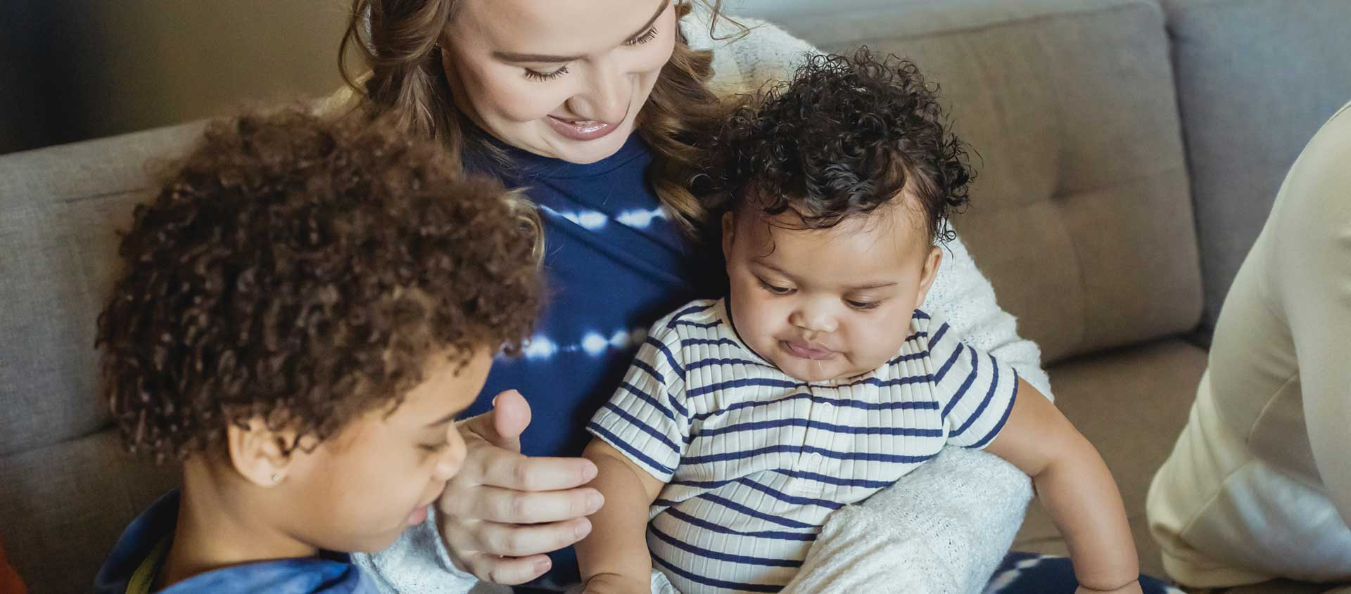 A Caucasian mother holds and Black infant while her Black daughter shows some play food to the infant.