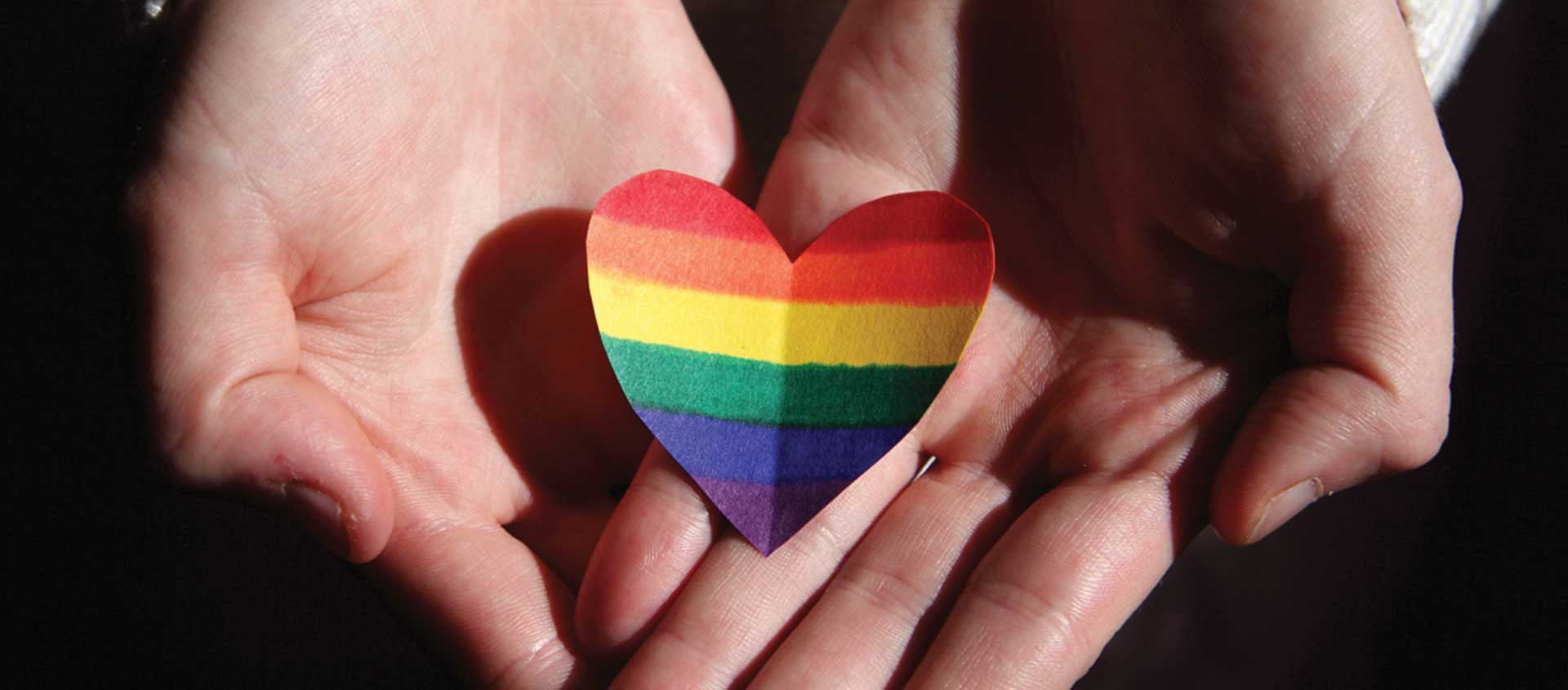 A pair of adult, Caucasian hands hold a paper heart cutout that has been colored with a rainbow. 