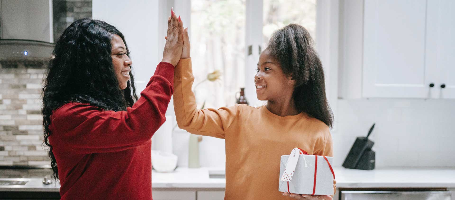 A Black American mother and daughter exchange a high-5 while the daughter holds a holiday present.