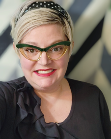 Candy Freed is a Caucasian woman with a blonde pixie cut. She is wearing a black top and matching headband with retro-style glasses