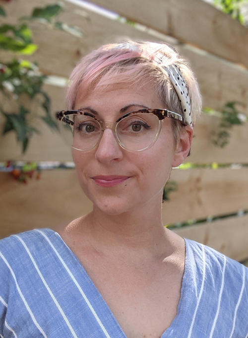 A Caucasian woman with short blonde hair that has pink highlights. She is wearing glasses and a blue top.