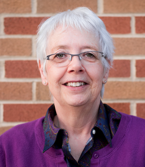 A Caucasian woman with short, silver hair and glasses, wearing a purple, button-down top.